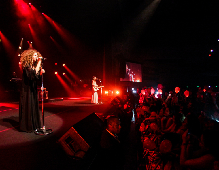 Cantora da dupla Anavitória se apresenta no palco iluminado por luzes vermelhas, enquanto o público assiste com balões vermelhos em mãos em um show ao vivo.