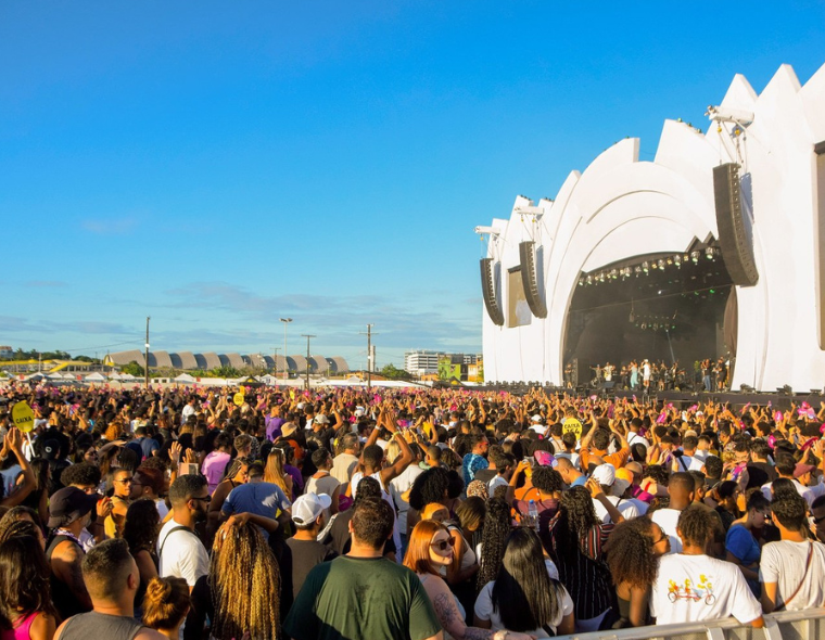 Multidão de pessoas em festival ao ar livre durante o dia, assistindo a apresentação em um grande palco branco com estrutura moderna, sob céu azul.
