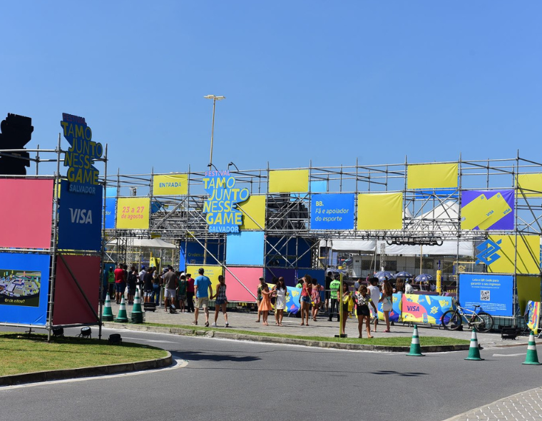 Entrada e estrutura do Festival Tamo Junto Nesse Game em Salvador, com placas coloridas de divulgação, público em fila e céu claro.