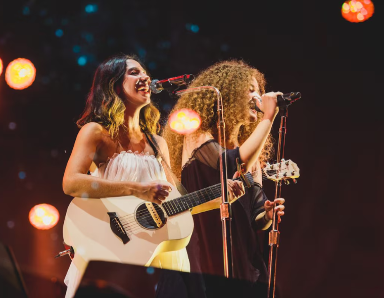 Dupla AnaVitória cantando ao vivo com Ana Caetano tocando violão e Vitória Falcão no microfone durante show com iluminação de palco em Maringá.