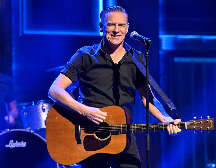 Bryan Adams cantando ao vivo com guitarra acústica durante show da turnê Roll With Punches, em palco com iluminação azul.