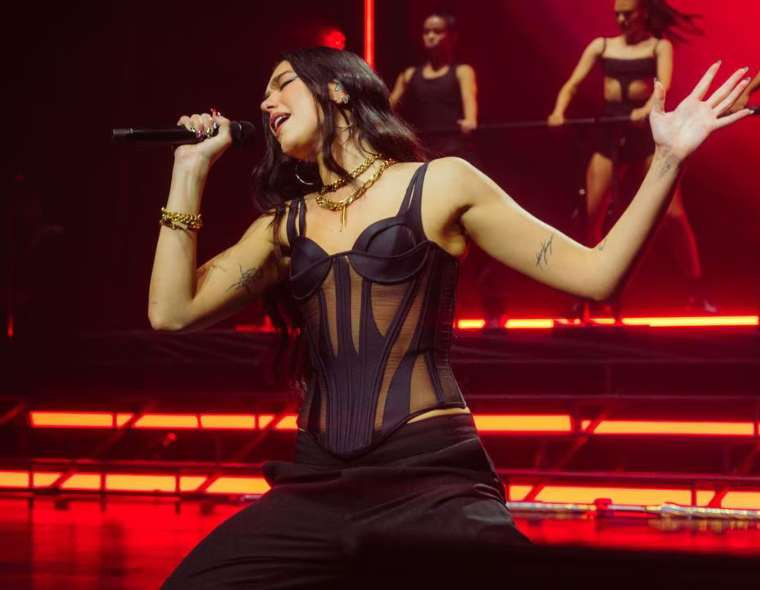 Cantora Dua Lipa performando ao vivo no palco, vestindo top preto estilo corselet e calça preta, segurando microfone com uma mão e com a outra levantada, com bailarinas ao fundo e iluminação vermelha.