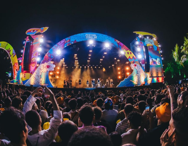 Grande público assistindo a apresentação no palco principal do Festival Zepelim à noite, com iluminação colorida, palco decorado com arte vibrante e artistas no palco, no Marina Park Hotel em Fortaleza.