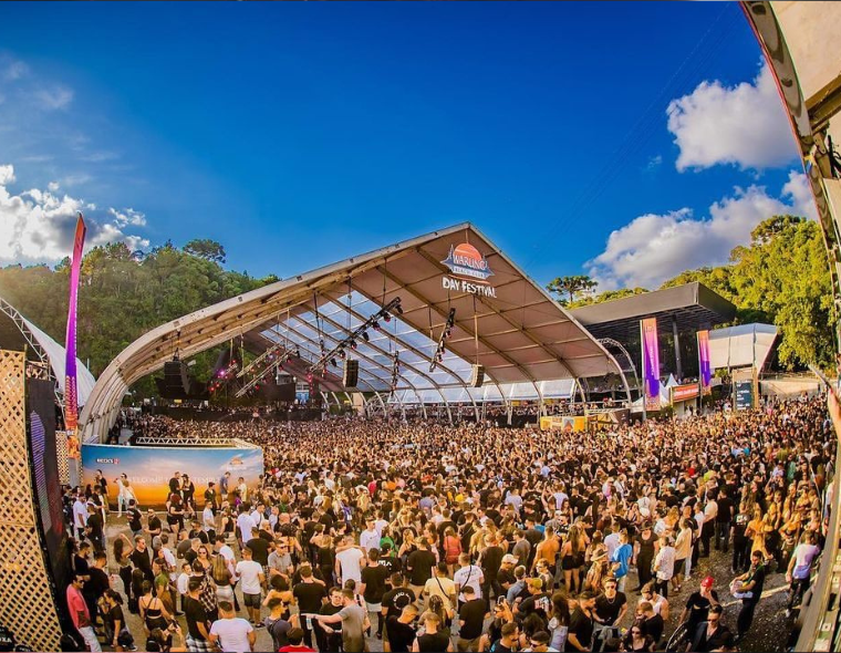 Grande público reunido no palco principal do Warung Day Festival durante evento em espaço aberto com estrutura coberta, em ambiente ensolarado com céu azul.