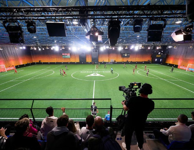 Partida de futebol 7 sendo transmitida ao vivo em arena indoor com torcida assistindo das arquibancadas e câmera profissional registrando o jogo.