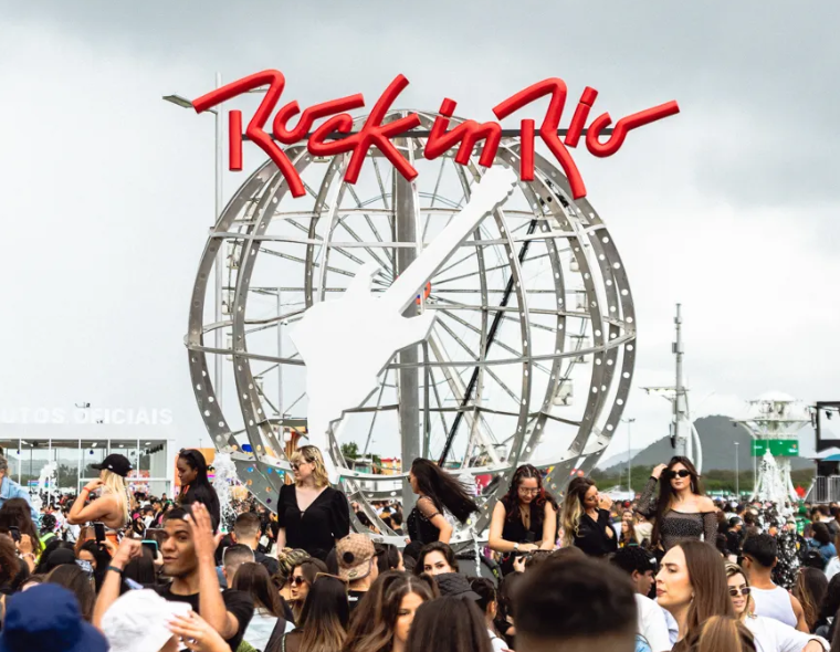 Multidão de pessoas na entrada da Cidade do Rock com logo vermelho do Rock in Rio e escultura metálica de guitarra ao fundo em dia nublado.