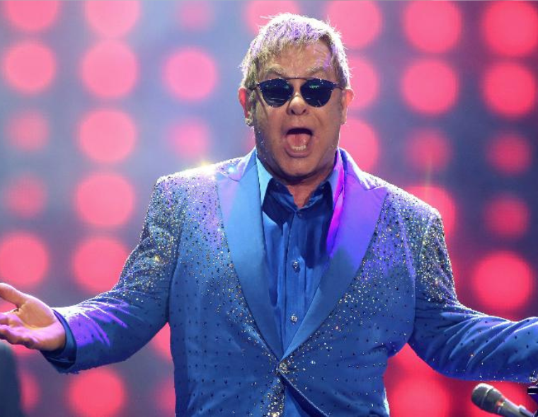 "Cantor (Elton John) vestido com terno azul brilhante e camisa azul, usando óculos escuros, performando no palco com luzes vermelhas desfocadas ao fundo.