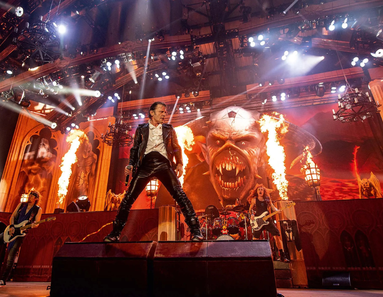 Show do Iron Maiden com o vocalista no centro do palco, banda tocando ao fundo e efeitos de fogo com imagem do mascote Eddie projetada na tela gigante.