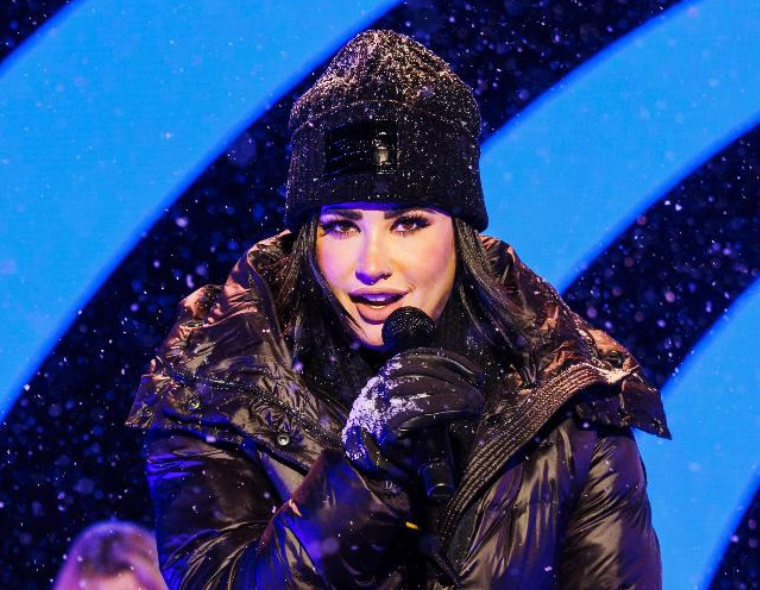 Demi Lovato se apresentando ao vivo com microfone, vestindo casaco preto e gorro, durante show em cenário com neve.