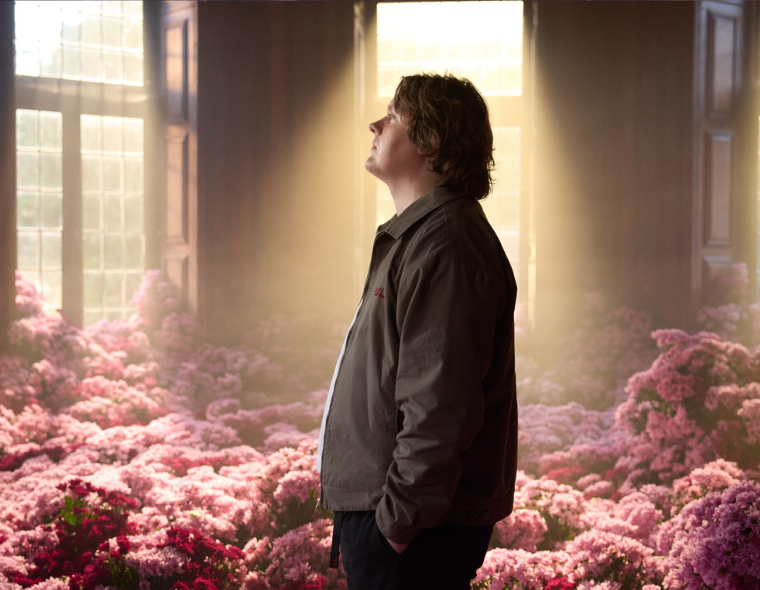Retrato de Lewis Capaldi em perfil, em ambiente iluminado por luz natural, rodeado por muitas flores rosas, transmitindo uma atmosfera introspectiva e serena.