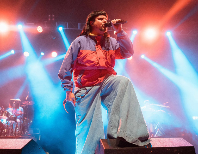 Oliver Tree cantando ao vivo em show com luzes coloridas de palco, vestindo jaqueta colorida e calça larga durante turnê Love You Madly Hate You Badly em São Paulo.