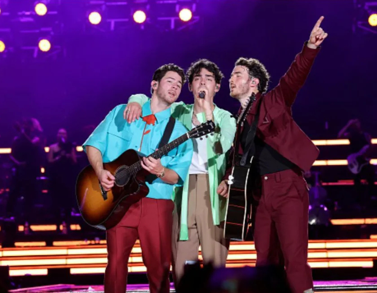 Jonas Brothers cantando juntos no palco durante show ao vivo, com instrumentos e iluminação vibrante