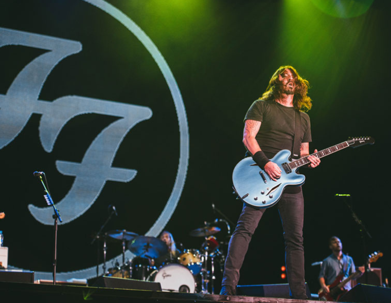 Dave Grohl, vocalista e guitarrista do Foo Fighters, tocando guitarra azul no palco durante show ao vivo com o logo da banda ao fundo.