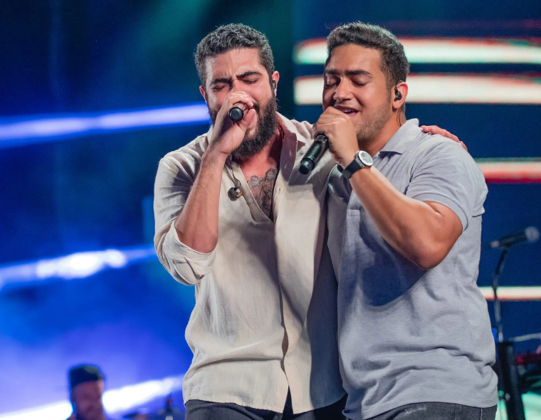 Henrique e Juliano, cantando juntos em um show, cada um segurando um microfone, com luzes coloridas ao fundo.