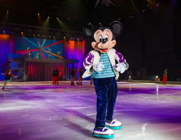 Mickey Mouse em apresentação no Disney On Ice SP, patinando no gelo com figurino moderno e iluminação colorida no palco.