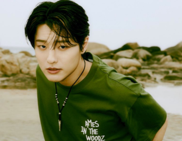 Close-up focado do jovem artista coreano Cho Seung-youn (WOODZ) com cabelos pretos penteados, em uma praia rochosa. Ele veste uma camiseta verde-oliva com o gráfico de texto branco 'AMES IN THE WOODZ' e um colar de contas com pingente. Luz natural difusa.